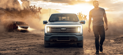 A man walking beside a blue 2023 Ford F-150 Lightning, while a red 2023 F-150 Lightning drives in the background and stirs up clouds of dust.