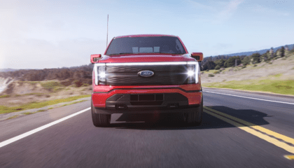 A red 2023 Ford F-150 Lightning shown from the front and driving down a coastal road during the day.
