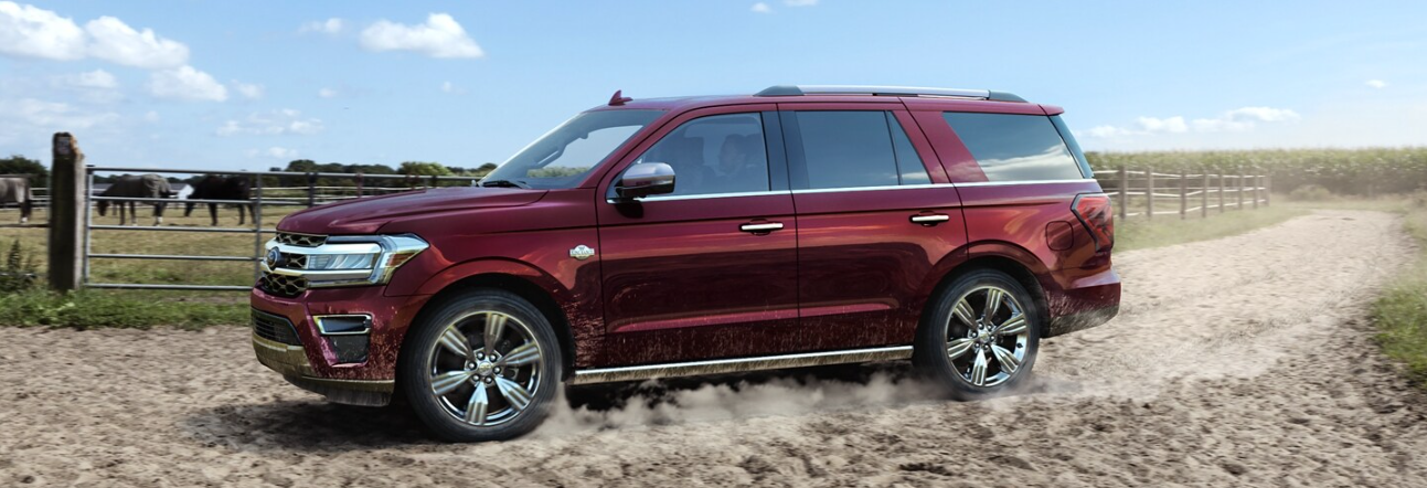 A red 2024 Ford Expedition King Ranch driving down a dirt road, with dust trailing its tires and cows in a field shown in the background.