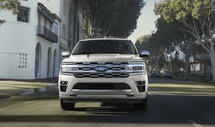 A white 2024 Ford Expedition King Ranch driving down a city street, with buildings and trees in the background.