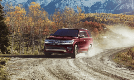 A red 2024 Ford Expedition King Ranch driving down a dirt road, with a dust cloud, trees, and mountains seen behind it.