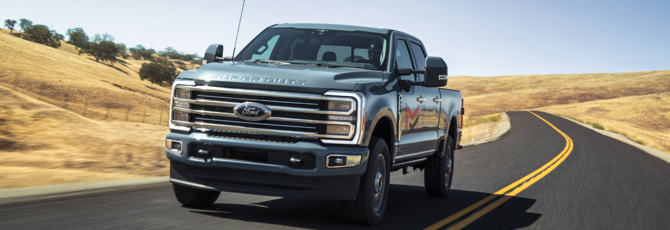 A silver 2024 Ford F-350 driving down a remote paved road, with grass and shrubs surrounding it.