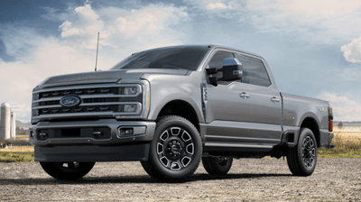 A silver 2024 Ford F-350 Platinum parked on a gravel road, with a field and silo visible in the background.