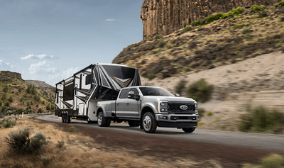 A silver 2024 Ford F-350 Limited pulling an  RV trailer down a desert road, with cliffs and shrubs surrounding it.