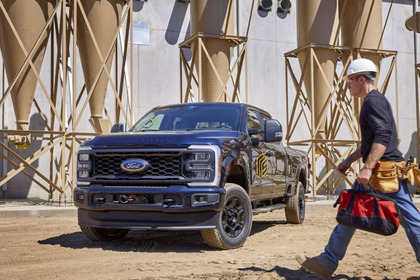 A black 2024 Ford F-350 parked at a work-site, with man in a hard-hat walking in front of it and various silos seen in the background.