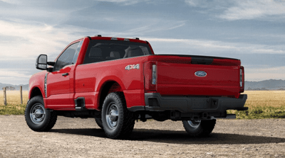 A red 2024 Ford F-350 XL parked on a gravel road, with a field, wire fence, and mountains visible in the background.