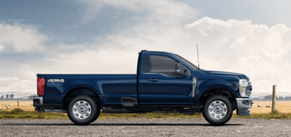 A blue 2024 Ford F-350 XLT shown in profile while parked on a gravel road. A wire fence and field are visible in the background.