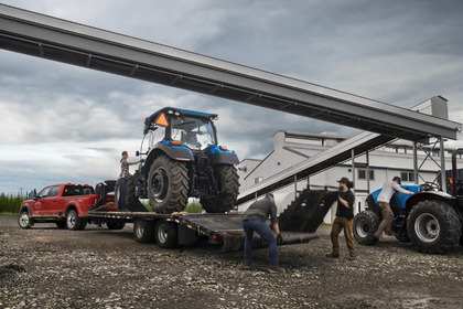 A red 2024 Ford F-350 shown hitched to a trailer carrying a tractor, with various other farm equipment and workers around it.