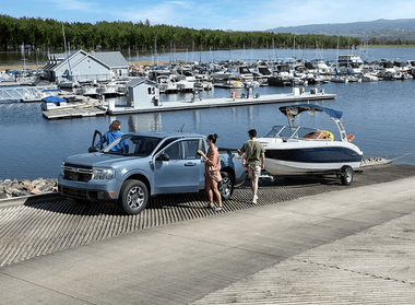 A blue 2024 Ford Maverick parked by a lake, with a boat hitched to it. Various other boats and docks are visible in the background.