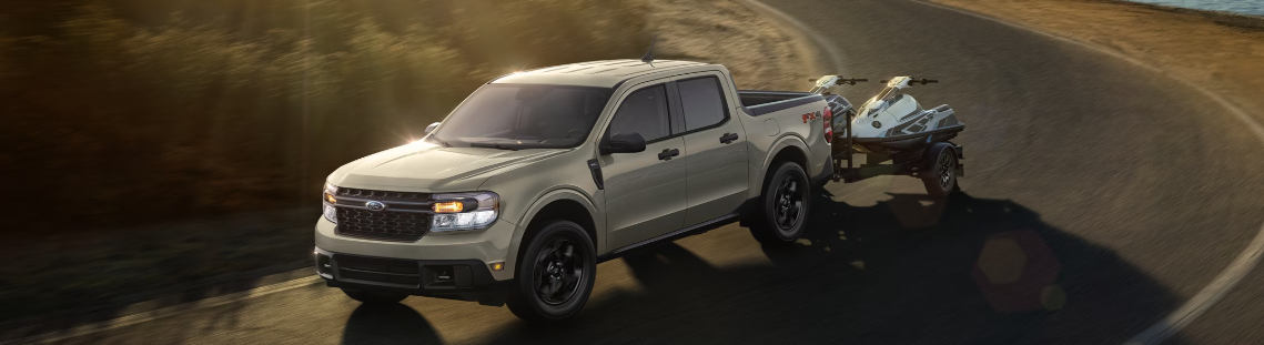 A silver 2024 Ford Maverick pulling an ATV down a winding road.