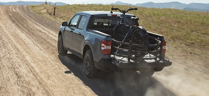 A black 2024 Ford Maverick driving down a dirt road, with a bike in its bed and dust trails following it.