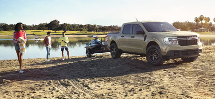 A silver 2024 Ford Maverick parked on a beach, with a lake in the background and people standing beside it.