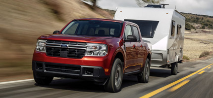 A red 2024 Ford Maverick pulling a white trailer down a road, with shrubs and sand in the background.