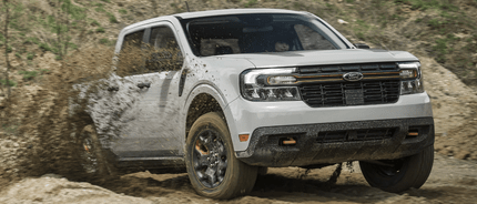 A white 2024 Ford Maverick equipped with the Tremor Package and driving down a dirt road, with mud being slung around it.