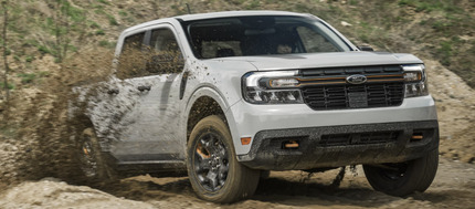 A white 2024 Ford Maverick driving down a dirt road, with mud slinging up around it and mountains visible in the backround.