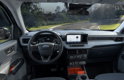 Interior view of the 2024 Ford Maverick XLT, with the steering wheel, dashboard, and eight-inch touchscreen displayed. A tree-lined road is visible through the windshield.