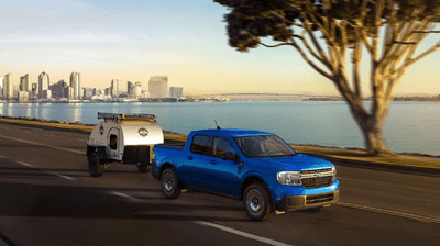 A blue 2024 Ford Maverick Lariat driving down a road while pulling a trailer. A city and lake are in the background.