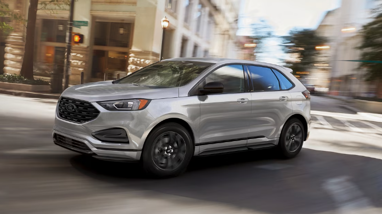 A silver 2024 Ford Edge driving down a city road, with buildings and street lights in the background.