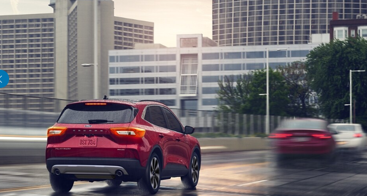 A red 2024 Ford Escape Platinum driving down a wet city street, with buildings and blurred cars shown in the background.