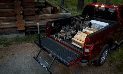 Rear view of a red 2023 Ford F-150 Limited, showcasing the power tailgate, available step feature, and work surface. Various cargo items are in the bed, and the truck is parked beside a cabin.