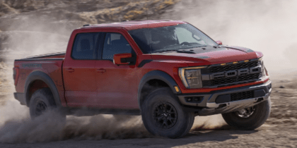 A red 2023 Ford F-150 Raptor driving on a dirt road, with dust clouds rising up around it.