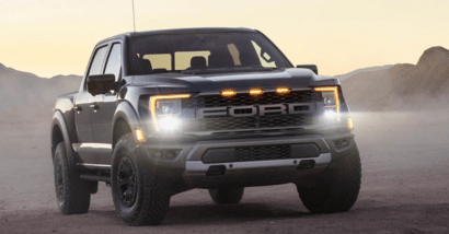 A black 2023 Ford F-150 Raptor parked in a desert-region at dusk, with its headlights activated and rock formations in the background.