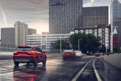 A red 2024 Ford Escape Platinum driving down a wet city street, with buildings and blurred cars shown in the background.