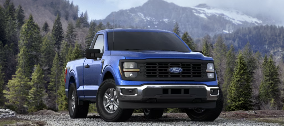 A blue 2024 Ford F-150 XL parked on a gravel road, with trees and snow-capped mountains visible in the distance.