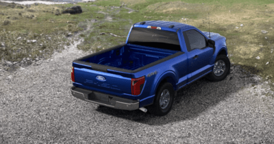 Rear view of a blue 2024 Ford F-150 XL parked on a gravel path, with grass visible in the distance.