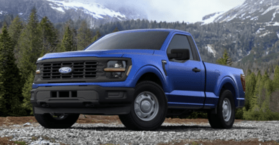 A blue 2024 Ford F-150 XL parked on a gravel path, with trees and mountains in the background.