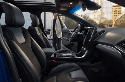 Interior view of the 2024 Ford Edge ST, with its leather seats, dashboard, and steering wheel displayed. A man in a white blazer is entering the car from the side.