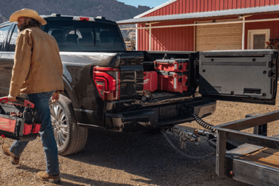 Rear view of a black 2024 Ford F-150 King Ranch, with its tailgate open and cargo visible in the bed. Beside it a man in a cowboy hat is carrying a toolbox.