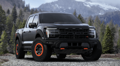 A black 2024 Ford F-150 Raptor with orange accents parked on a gravel road. Mountains and a forest are in the background.