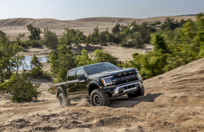 A black 2024 Ford F-150 Raptor driving up a steep sandy hill, with trees, shrubs, and mountains in the background.