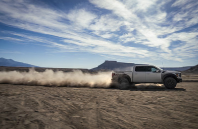 A white 2024 Ford F-150 Raptor driving across the desert, with a dust trail visible behind it.