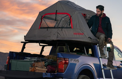 Rear view of a blue 2024 Ford F-150 STX, with a tent installed across the bed and a man climbing into the tent.