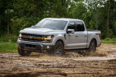 A silver 2024 Ford F-150 driving through mud, with trees shown in the background.