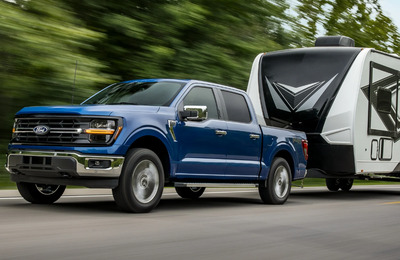 A blue 2024 Ford F-150 XLT pulling a black and white trailer down a tree-lined road.