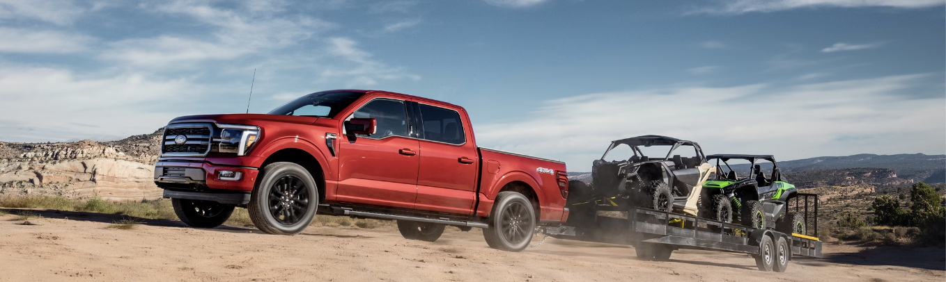 A red 2024 Ford F-150 parked in the desert, with a trailer hauling ATVs parked behind it and mountains visible in the distance.