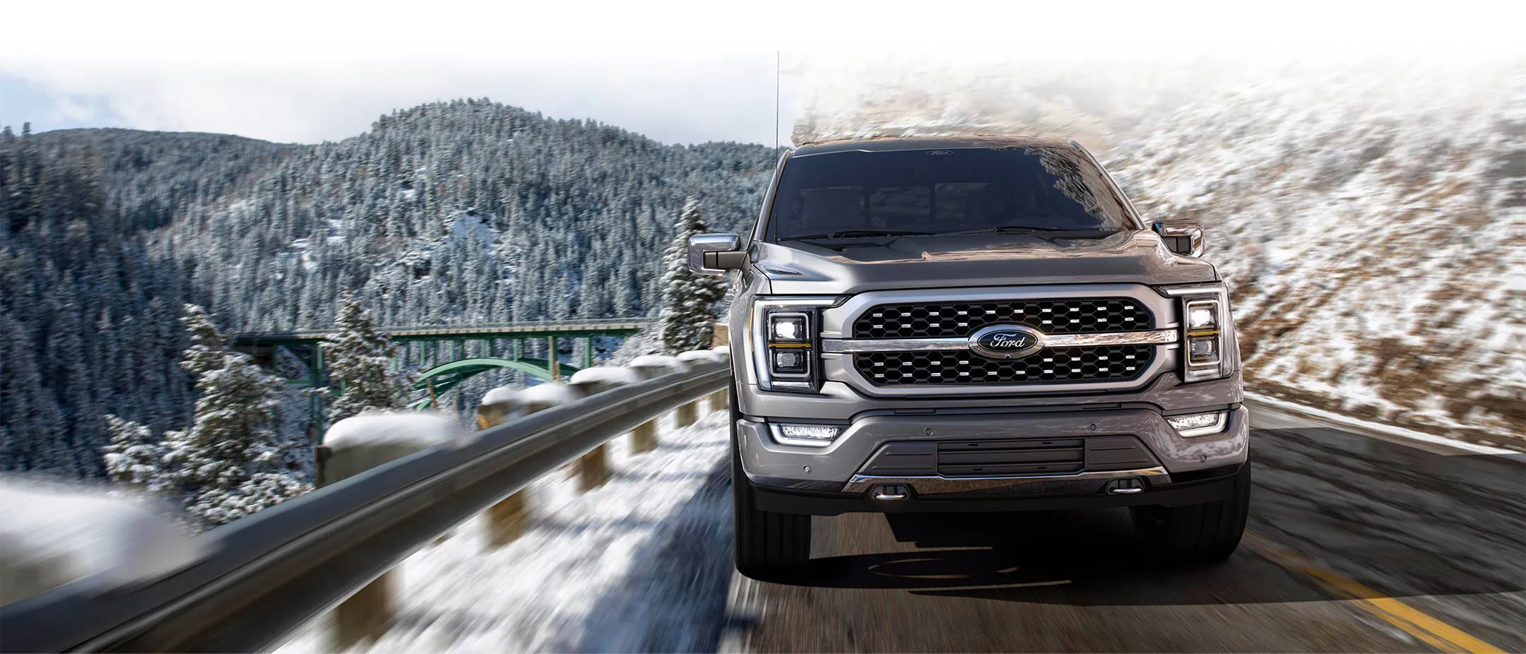 A silver 2023 Ford F-150 shown from the front and driving down a mountain road, with trees and snow in the background.
