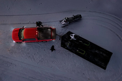Aerial view of a red 2023 Ford F-150 parked in the snow, with a trailer and snowmobile sat behind it. Two people also stand beside it, and the lights are all activated.