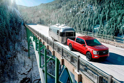 A red 2023 Ford F-150 pulling an Airstream trailer across a bridge, surrounded by trees and snow.