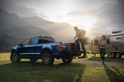 A blue 2023 Ford F-150 King Ranch parked in a field, with a man climbing into the rear and a woman standing beside it. Mountains are visible in the distance, and an Airstream is parked beside it.