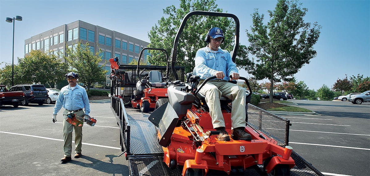 A man driving on a Husqvarna riding lawn mower off the back of a trailer, while another carries a trimmer. A commercial building and parking lot are seen in the background.