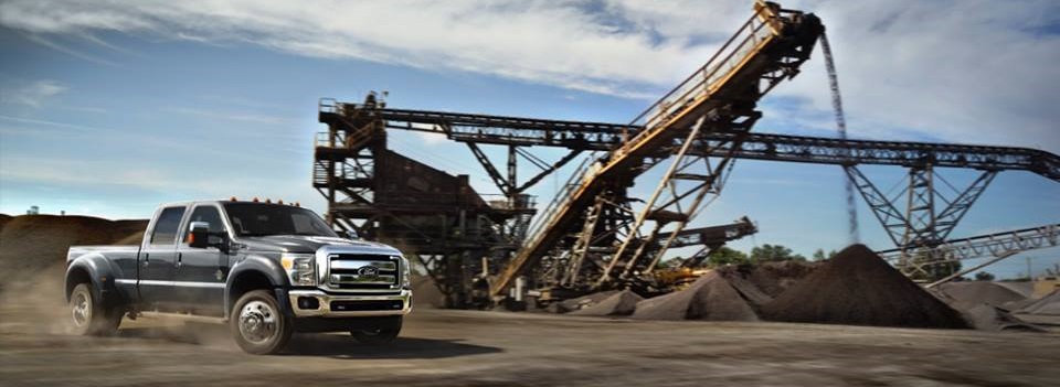 A dark grey Ford Super Duty parked at an industrial site, with cranes, piles of dirt, and other materials visible in the background.