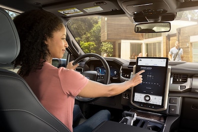 A woman sitting inside the 2022 F-150 Lightning and using the touchscreen. Outside, a man is seen walking toward the truck from the house.